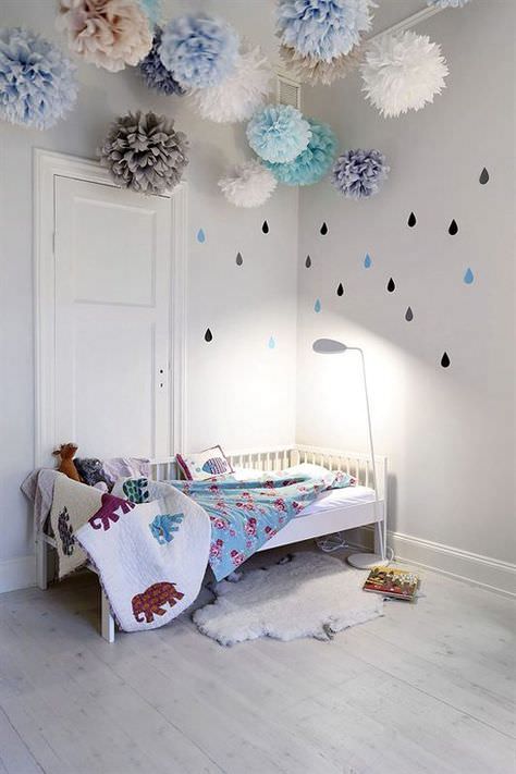اتاق خواب کودک که سقف آن با پام پام های کاغذی دکور شده است و دارای دیوار و تخت سفید رنگ می باشد